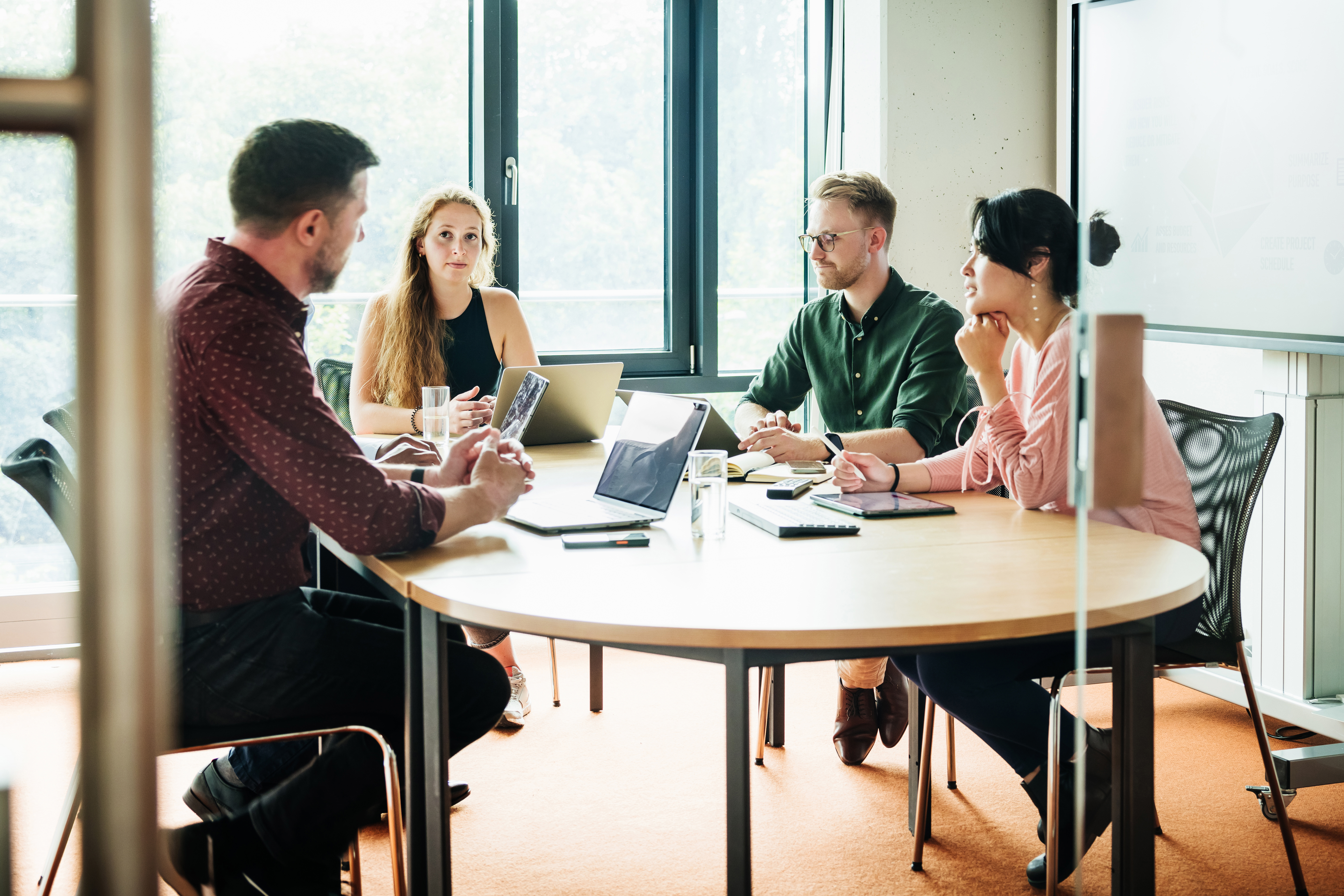 Vier Menschen sitzen in einer konzentrierten Arbeitssituation an einem runden Tisch in einem hellen Meetingraum, mit Notizen und digitalen Endgeräten in den Händen.