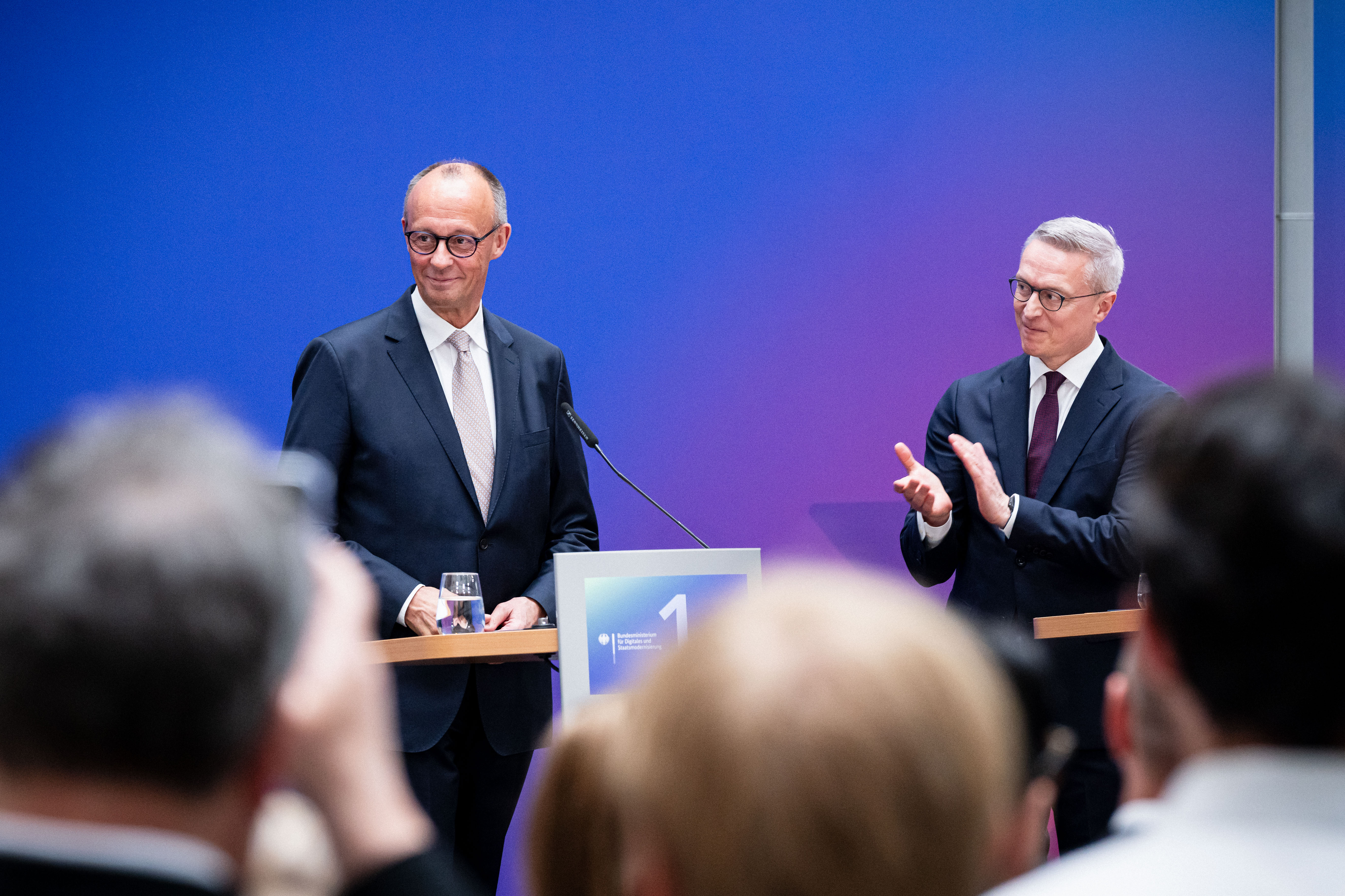 Friedrich Merz und Karsten Wildberger stehen an Rednerpulten auf einer Bühne, Wildberger applaudiert. Im Vordergrund ist unscharf das Publikum zu erkennen.