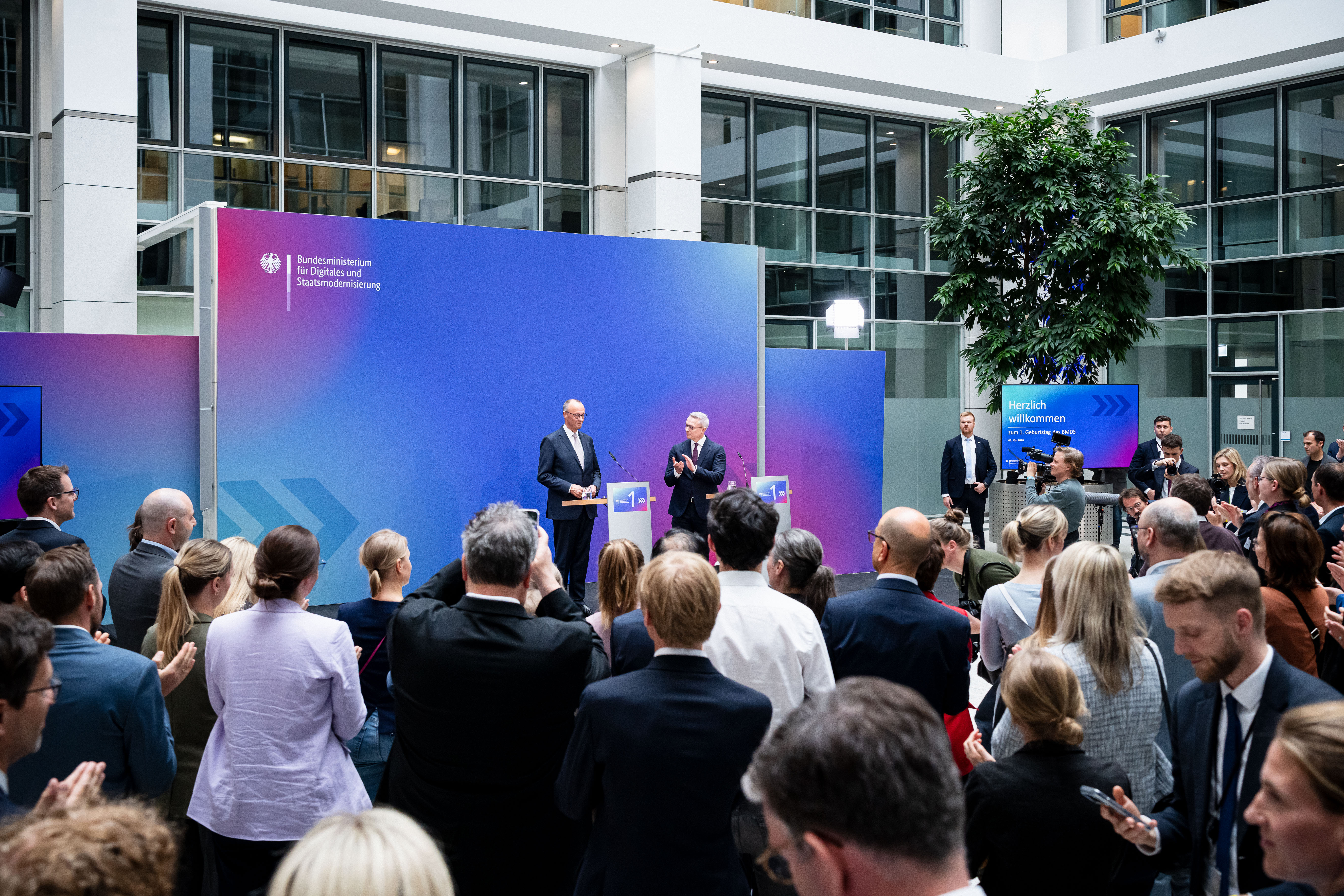 Friedrich Merz und Karsten Wildberger stehen an Rednerpulten auf einer Bühne, Wildberger applaudiert. Vor der Bühne stehen viele Menschen im Publikum.