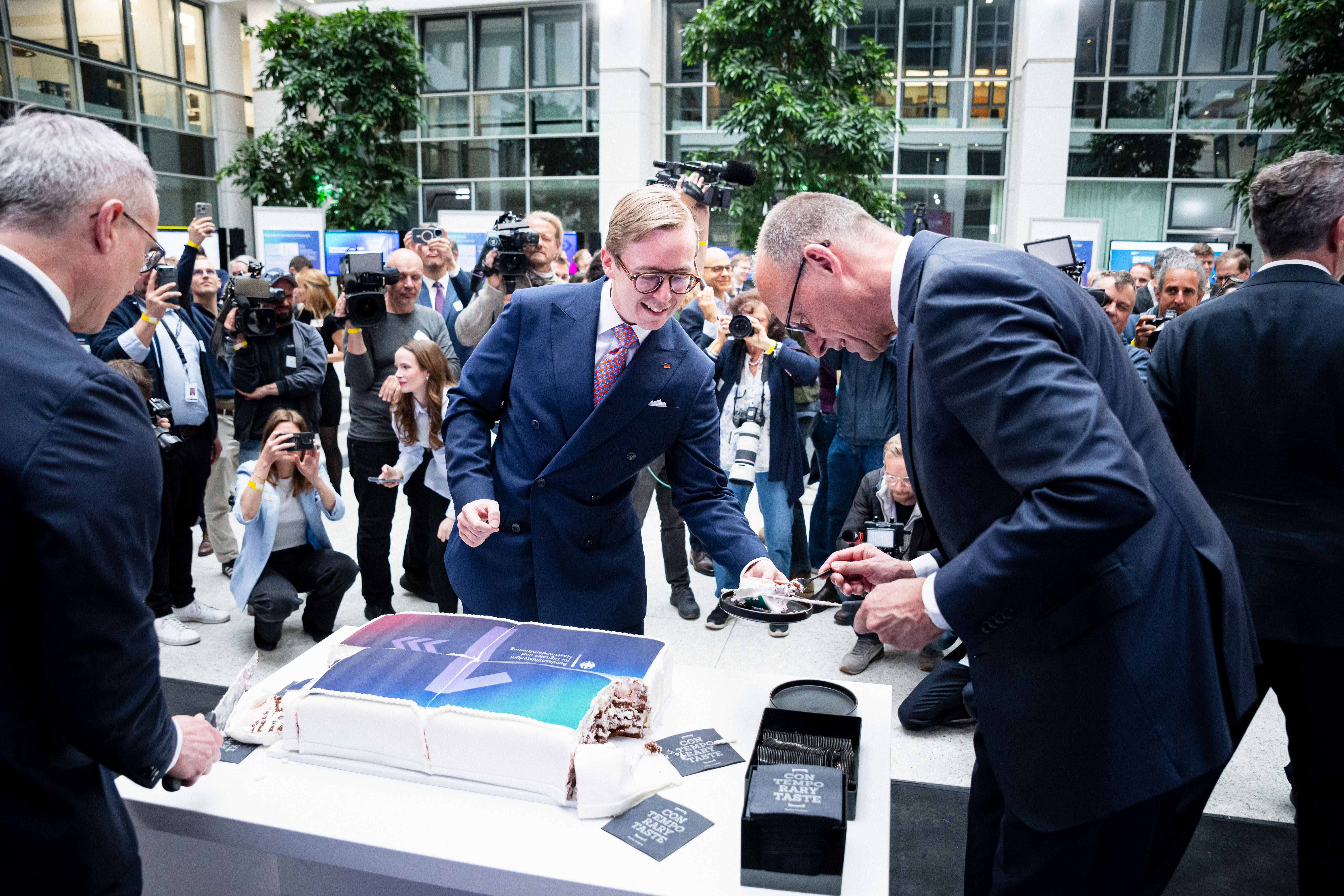 Friedrich Merz reicht Philipp Amthor einen Teller mit Kuchen. Im Hintergrund sind Fotografen zu sehen.