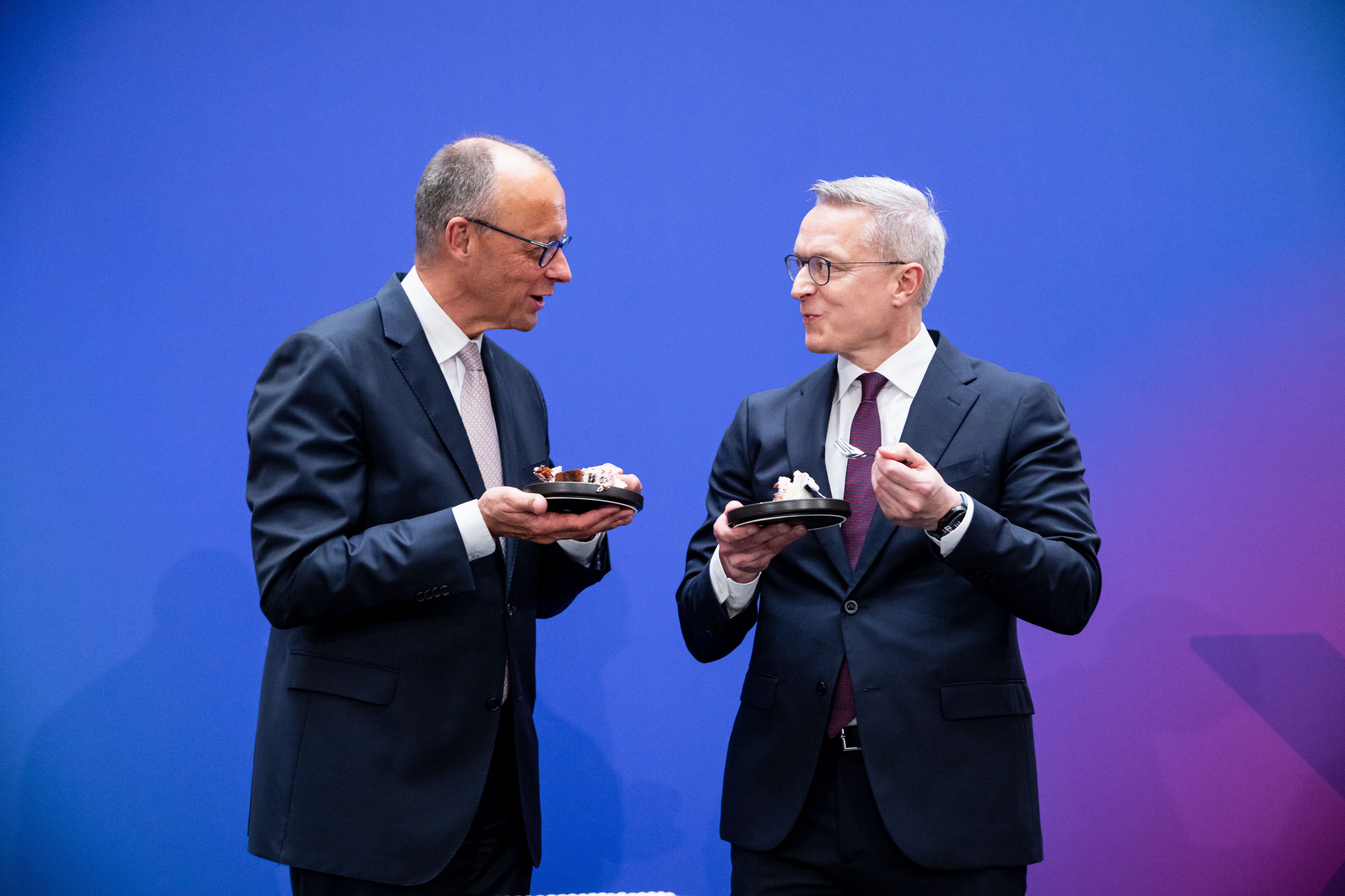 Friedrich Merz und Karsten Wildberger im Gespräch. Beide halten einen Teller mit Kuchen und eine Gabel in der Hand.