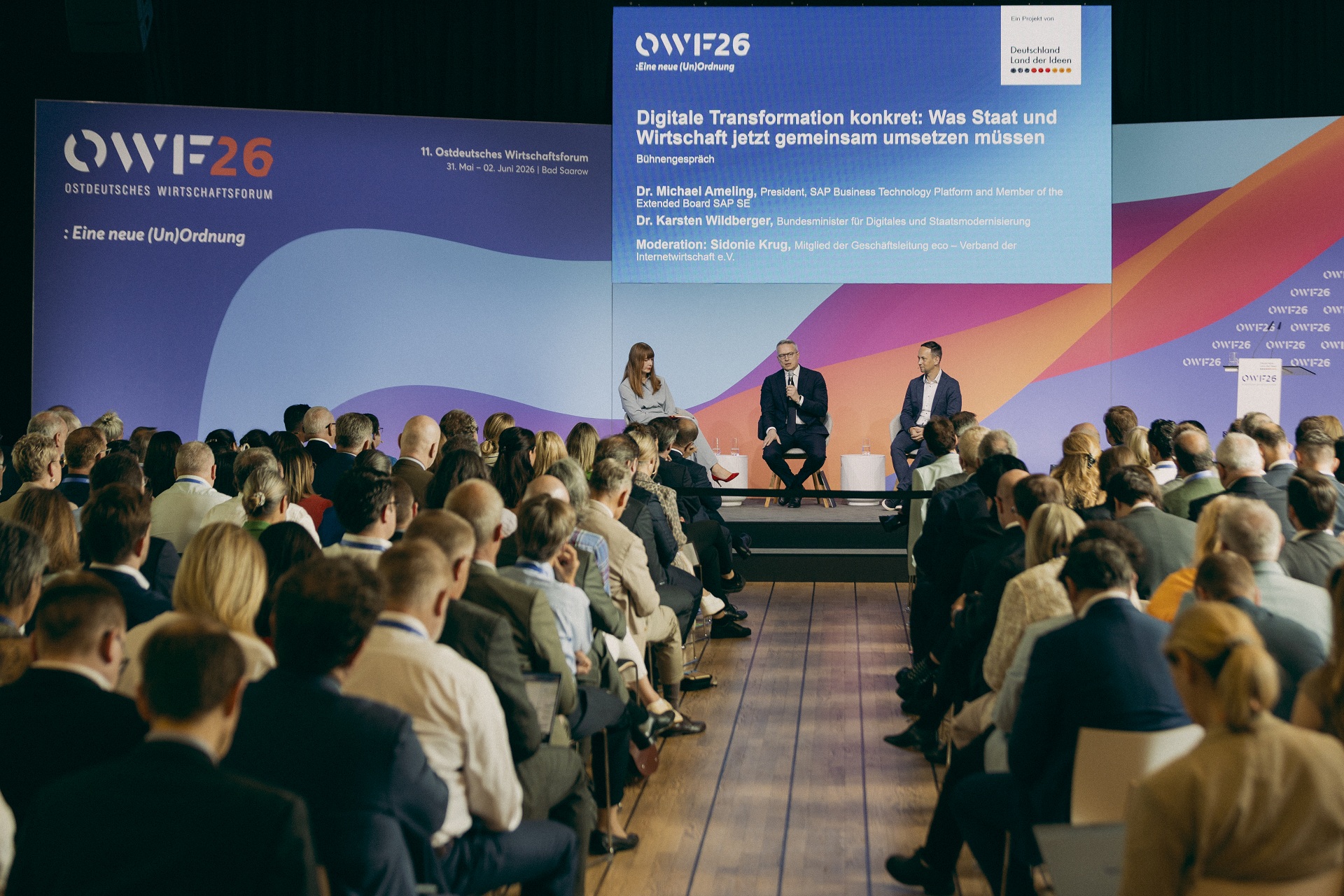 Auf einer Konferenzbühne vor großem Publikum sitzen drei Personen. Hinter ihnen ist eine Präsentationsfolie zum Ostdeutschen Wirtschaftsforum 2026 eingeblendet.