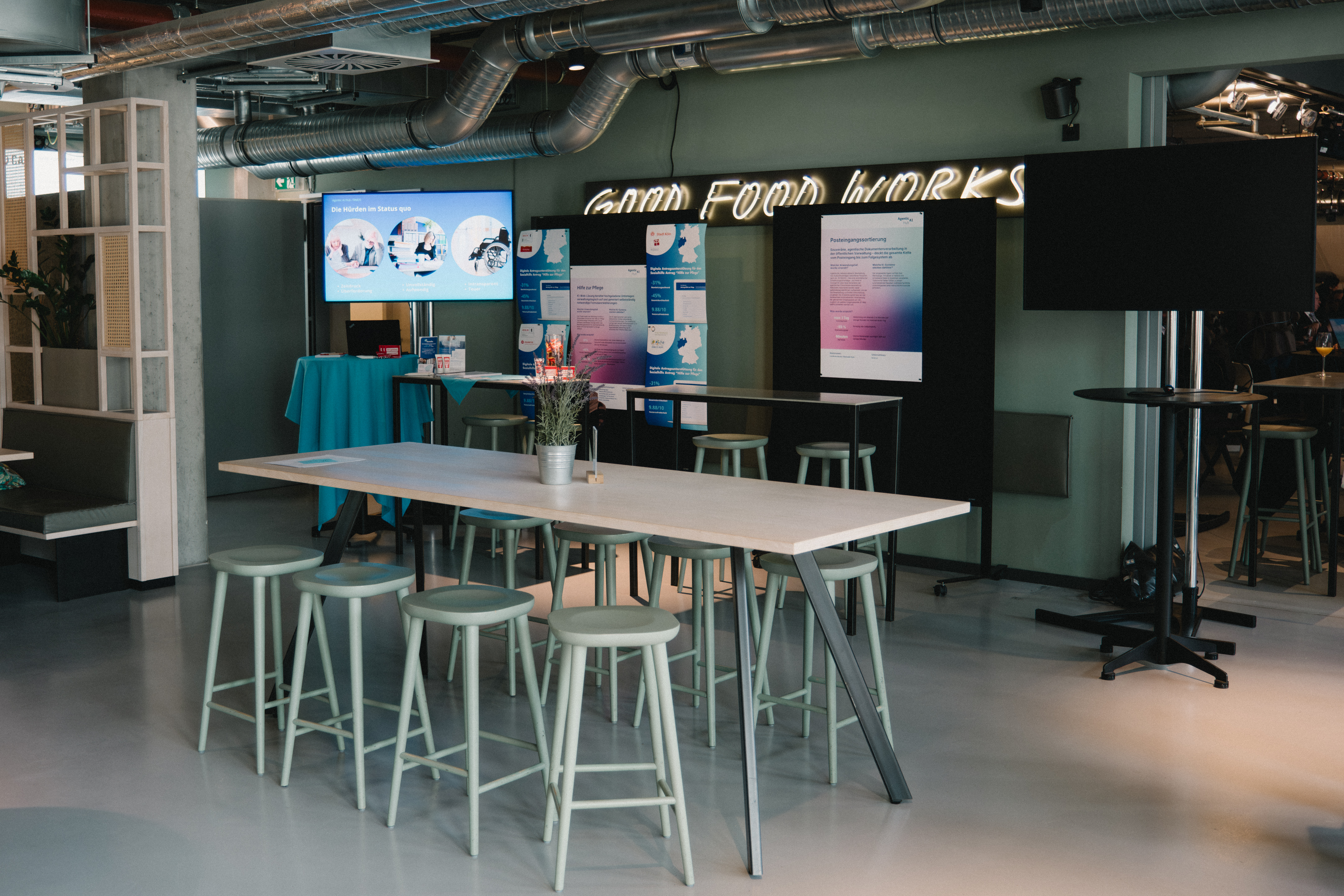 Ein moderner Veranstaltungs- oder Coworking-Raum mit einem großen Holztisch und hellgrünen Barhockern im Vordergrund. Im Hintergrund sind Infotafeln, Bildschirme und ein Neonschild mit der Aufschrift „GOOD FOOD WORKS“ zu sehen.
