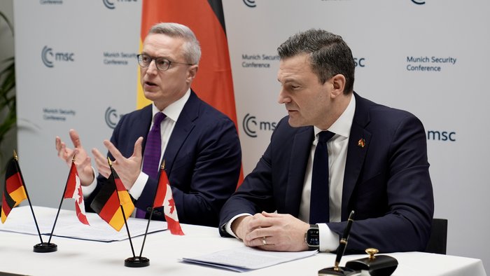 Der deutsche und der Kanadische Minister sitzen an einem Tisch. Auf dem Tisch stehen die deutsche und die kanadische Flagge.