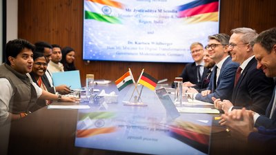 Fünf Personen sitzen links und fünf rechts an einem Tisch. Im Hintergrund ist ein großer Bildschirm an einer Wand.