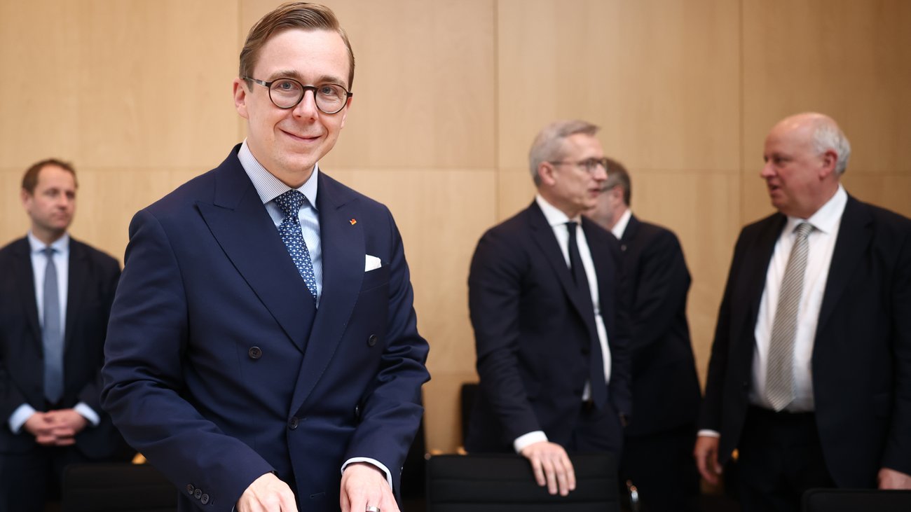Ein Mann im Anzug, vor ihm eine Aktentasche, steht an einem Sitzungstisch in einem Sitzungsraum und lächelt in die Kamera. Im Hintergrund weitere Ausschussmitglieder.