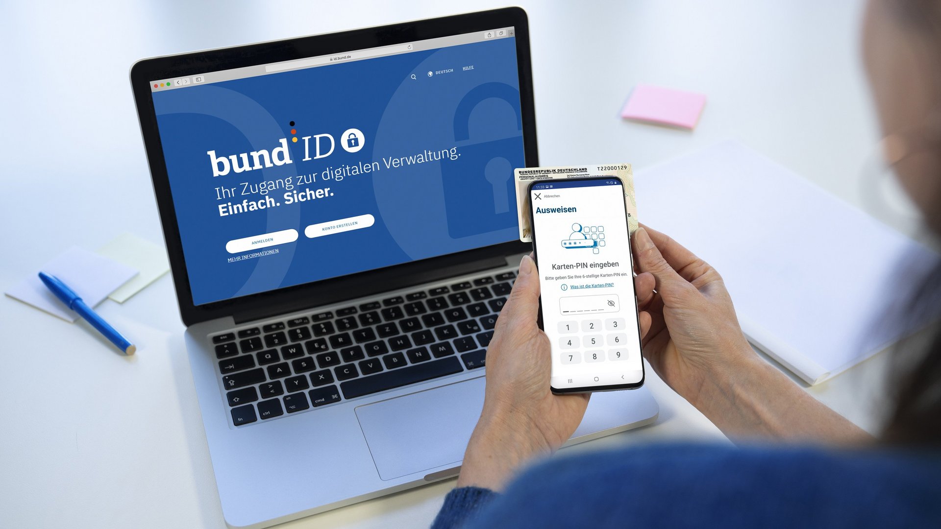 Eine Person sitzt vor dem Laptop, auf dem die Startseite der BundID aufgerufen wurde. Parallel hält sie ihren Personalausweis an die Rückseite ihres Smartphones, um sich nach Eingabe ihrer PIN mit ihrem Online-Ausweis online zu identifizieren.