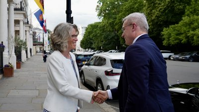 Ein Mann und eine Frau in Business-Kleidung geben sich auf einer londoner Straße die Hand zur Begrüßung. Im Hintergrund sieht man Flaggen an einer Häuserfassade.