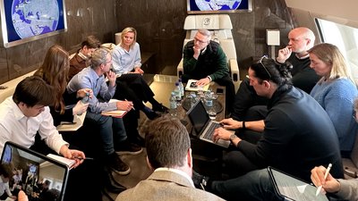 Mehrere Personen sitzen in einem Flugzeug um einen größeren Tisch und besprechen sich. Einige haben einen Laptop dabei.