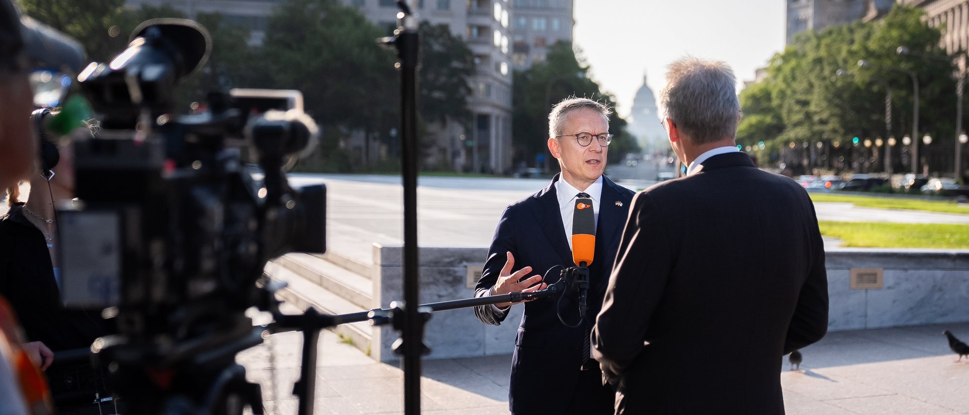 Zwei Personen in Businesskleidung, einer interviewt die andere Person. Am äußeren Rand sieht man Kameras. Ein Mikro wird ins Bild gehalten. 