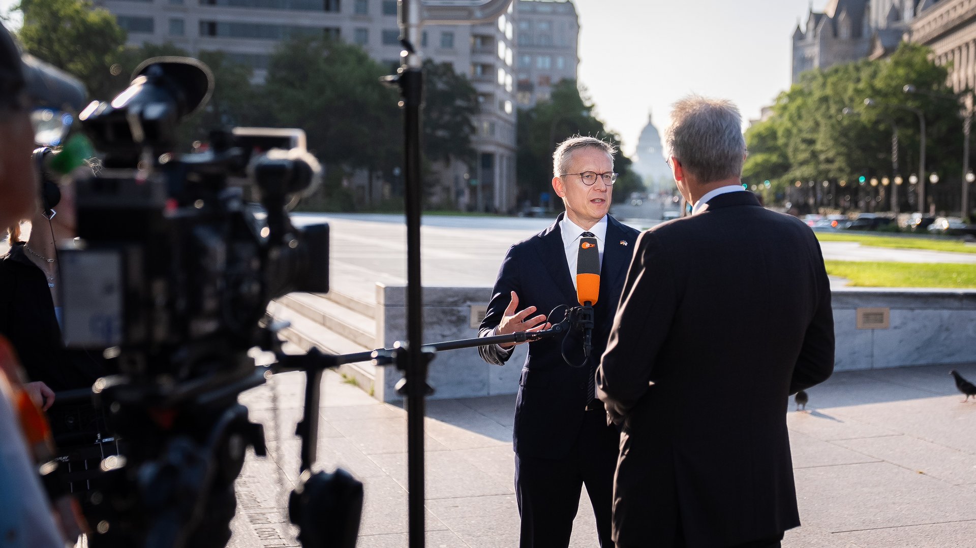 Zwei Personen in Businesskleidung, einer interviewt die andere Person. Am äußeren Rand sieht man Kameras. Ein Mikro wird ins Bild gehalten. 