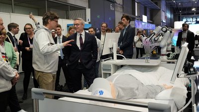 Der deutsche Bundesminister für Digitales und Staatsmodernisierung, Dr. Karsten Wildberger (Mitte, im dunklen Anzug), besucht eine Technologiedemonstration auf dem Mobile World Congress (MWC) 2026. Ein Experte (links, im hellgrauen Hoodie) erklärt ihm eine Anwendung im Bereich „Digital Health“. Im Vordergrund liegt eine medizinische Simulationspuppe in einem Patientenbett. Dahinter ist ein weißer, kollaborativer Roboterarm von Franka Robotics zu sehen, der für Aufgaben in der Pflege oder Medizin eingesetzt werden kann. Zahlreiche Messebesucher und Kamerateams beobachten die Szene in der Messehalle.