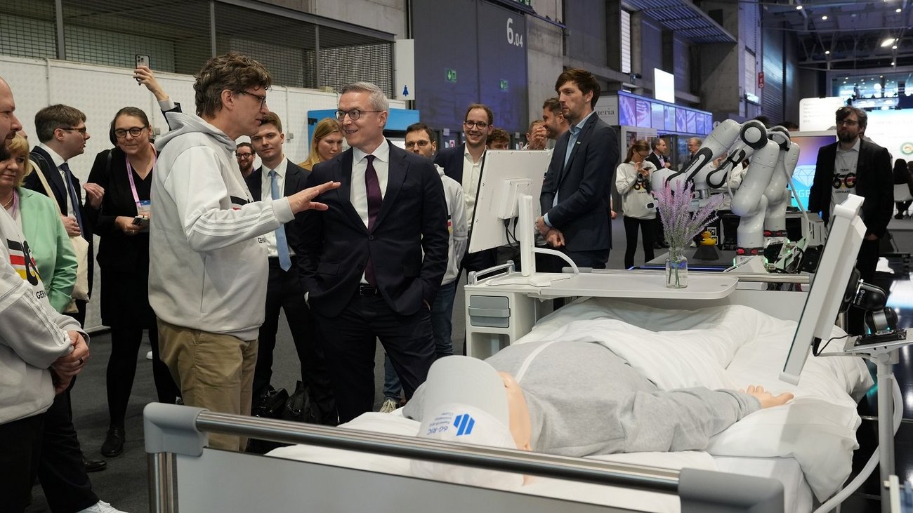 Der deutsche Bundesminister für Digitales und Staatsmodernisierung, Dr. Karsten Wildberger (Mitte, im dunklen Anzug), besucht eine Technologiedemonstration auf dem Mobile World Congress (MWC) 2026. Ein Experte (links, im hellgrauen Hoodie) erklärt ihm eine Anwendung im Bereich „Digital Health“. Im Vordergrund liegt eine medizinische Simulationspuppe in einem Patientenbett. Dahinter ist ein weißer, kollaborativer Roboterarm von Franka Robotics zu sehen, der für Aufgaben in der Pflege oder Medizin eingesetzt werden kann. Zahlreiche Messebesucher und Kamerateams beobachten die Szene in der Messehalle.