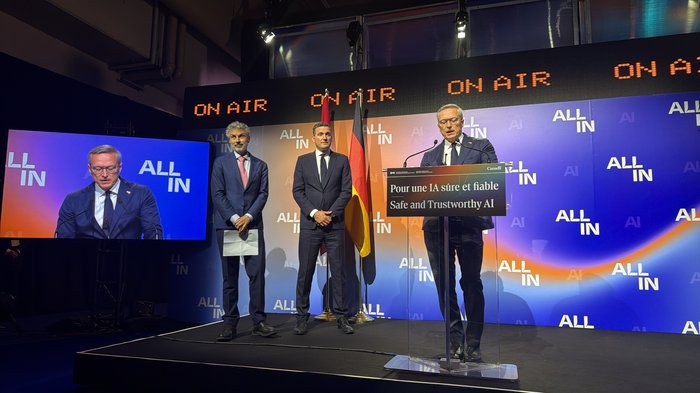 Drei Männer in Anzügen stehen auf der Bühne der ALL IN, einer redet am Rednerpult. Am Pult ist ein Schild befestigt, auf dem steht: "Pour une IA sure et fiable / Safe and Trustworthy AI!