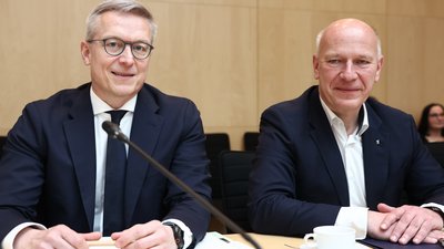 Zwei Männer in Anzügen sitzen am Tisch eines Sitzungsraums und blicken in die Kamera. Vor ihnen auf dem Tisch liegen Papiere. 