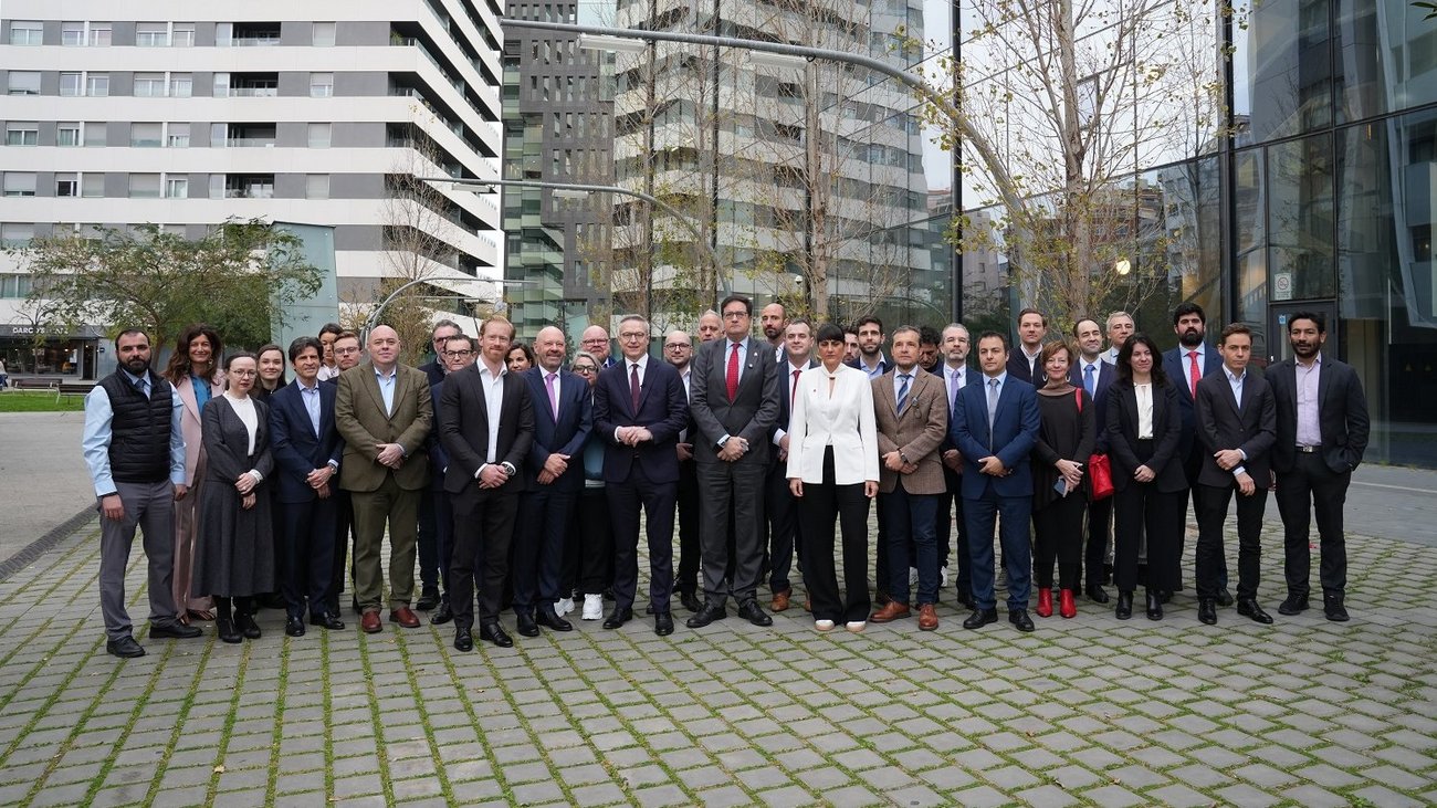 Eine große Gruppe von Männern und Frauen in Geschäftskleidung steht in zwei Reihen auf einem gepflasterten Platz vor modernen Glasgebäuden. Im Hintergrund sind hohe Wohn- und Bürohäuser sowie kahle Bäume zu sehen. Die Personen blicken direkt in die Kamera.