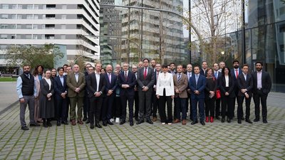 Eine große Gruppe von Männern und Frauen in Geschäftskleidung steht in zwei Reihen auf einem gepflasterten Platz vor modernen Glasgebäuden. Im Hintergrund sind hohe Wohn- und Bürohäuser sowie kahle Bäume zu sehen. Die Personen blicken direkt in die Kamera.