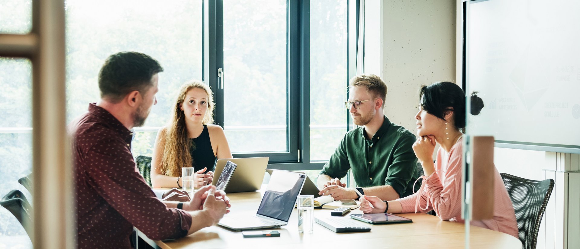 Vier Menschen sitzen in einer konzentrierten Arbeitssituation an einem runden Tisch in einem hellen Meetingraum, mit Notizen und digitalen Endgeräten in den Händen.