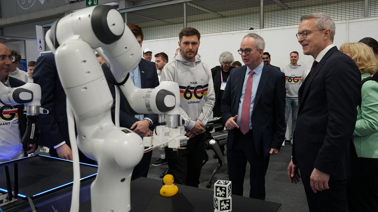 Ein weißer, gelenkiger Roboterarm steht im Fokus einer Technologiedemonstration auf einer Messe. Rechts im Bild betrachtet Karsten Wildberger im dunklen Anzug interessiert die Vorführung. In der Mitte steht ein junger Mann in einem grauen Hoodie mit der Aufschrift „Team Germany“, während im Hintergrund weitere Personen in Business-Kleidung die Szene beobachten. Vor dem Roboter auf einem schwarzen Tisch befinden sich eine gelbe Gummiente und zwei Würfel mit QR-Codes.