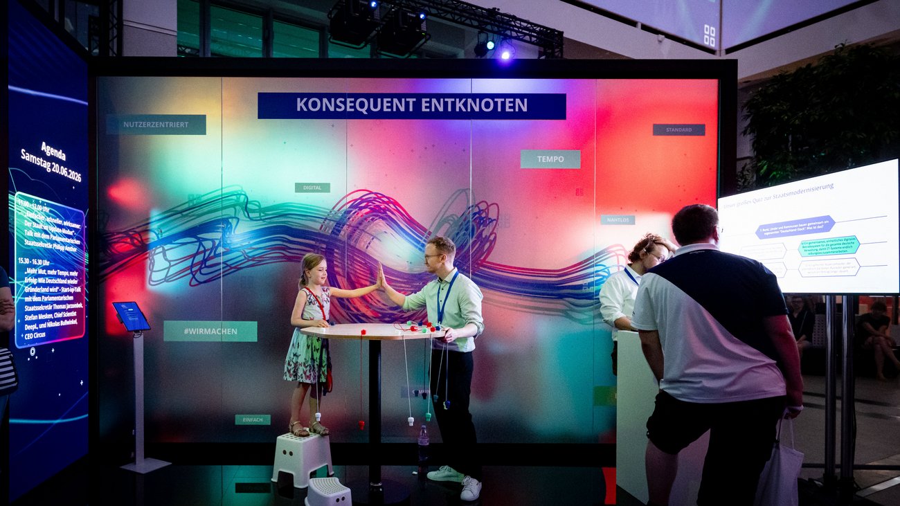 Mann und Kind vor einer großen Leinwand, darauf ein buntes Bild mit Schnüren und dem Slogan "Konsequent entknoten".