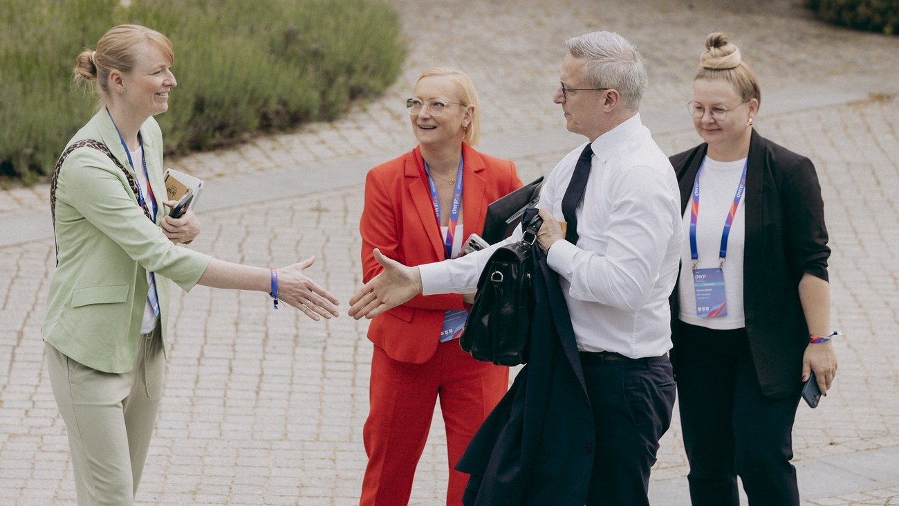 Karsten Wildberger reicht einer Frau in einem hellgrünen Blazer die Hand. Hinter ihnen stehen zwei weitere Frauen. Alle tragen Konferenz-Ausweise um den Hals.