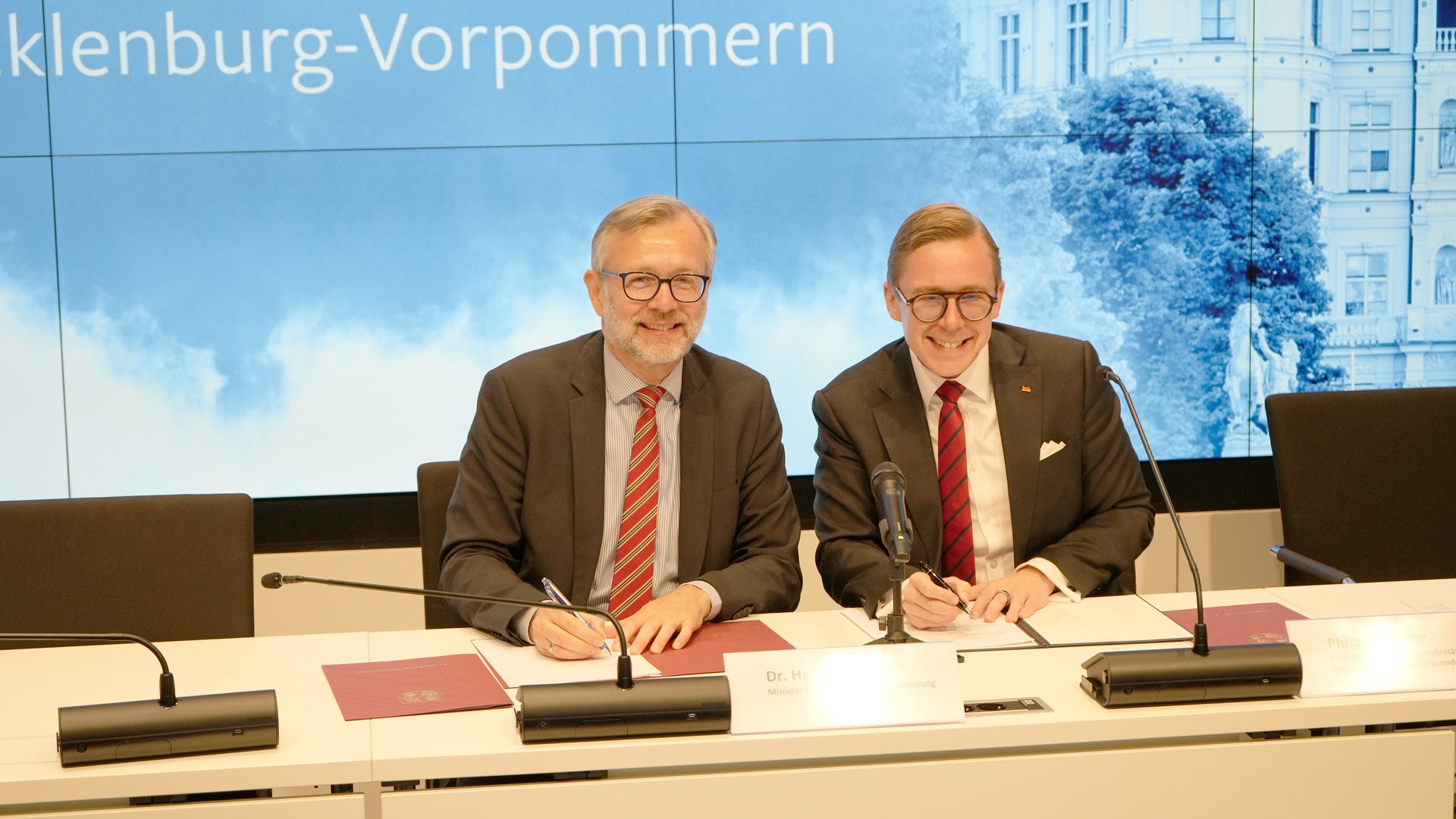 Zwei Männer in Anzügen sitzen an einem Tisch vor einer Pressewand und unterzeichnen Dokumente. Für das Foto schauen sie in die Kamera.