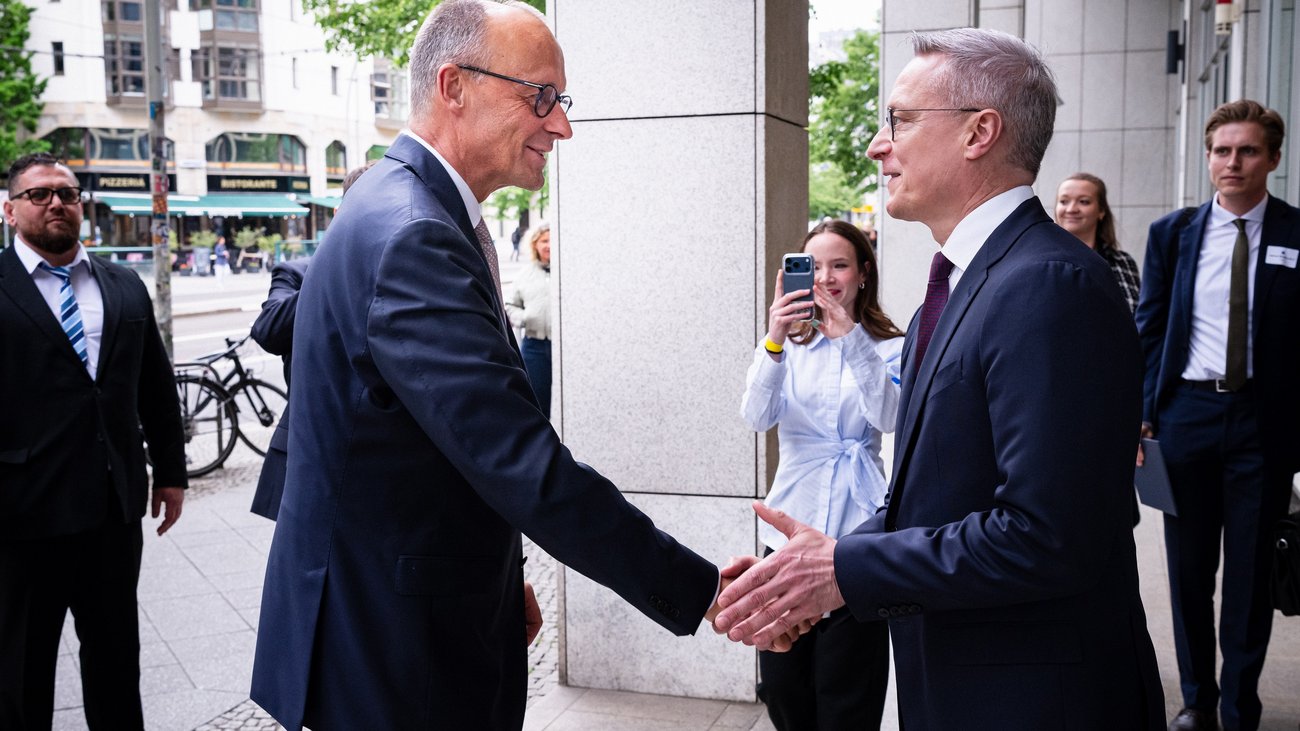 Nahaufnahme von Friedrich Merz und Karsten Wildberger (beide CDU), die sich lächelnd die Hand schütteln. Beide stehen im Profil vor einem modernen Gebäude. Im Hintergrund sind weitere Personen zu sehen, darunter eine junge Frau, die die Szene mit ihrem Smartphone fotografiert.