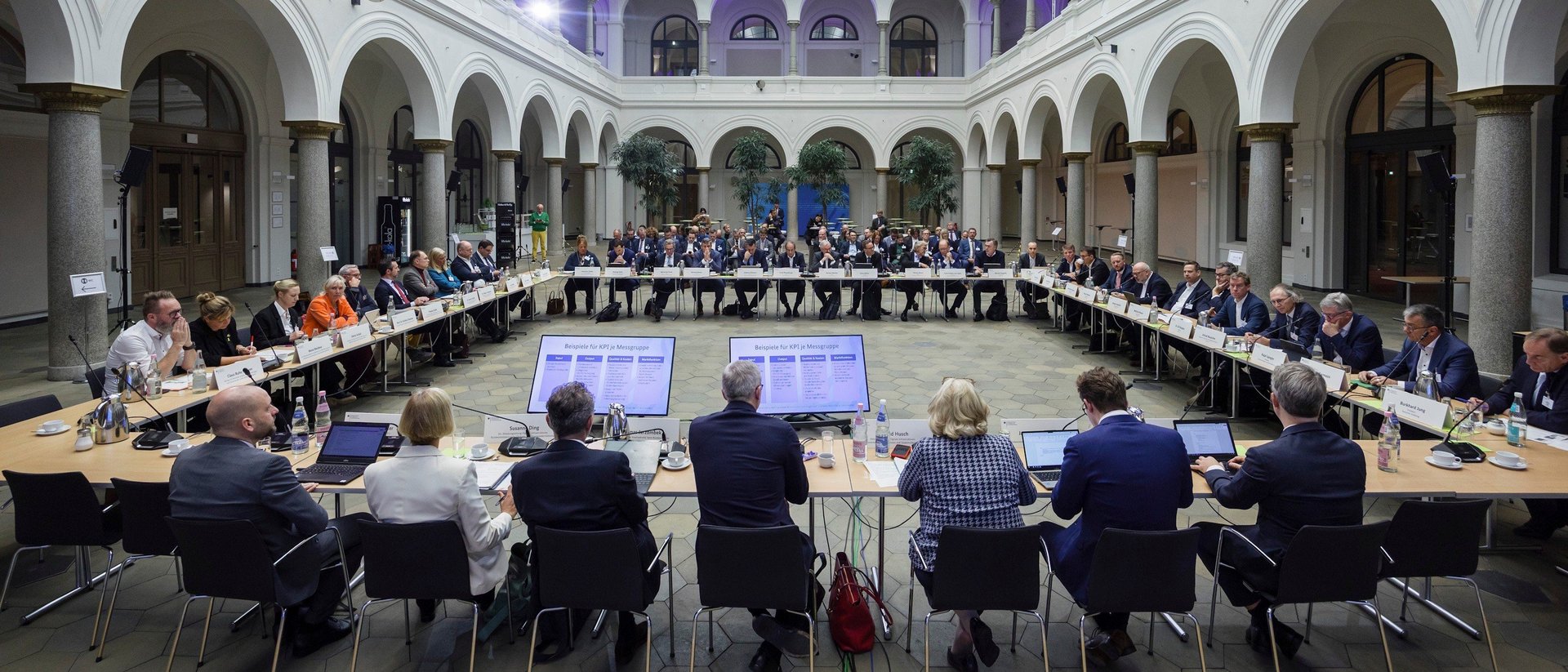 Blick in einen großen Saal mit Lichtauge und Arkaden, die den Raum umgeben. In einem Viereck sind Tische und Stühle aufgebaut, an denen die Anwesenden sitzen.