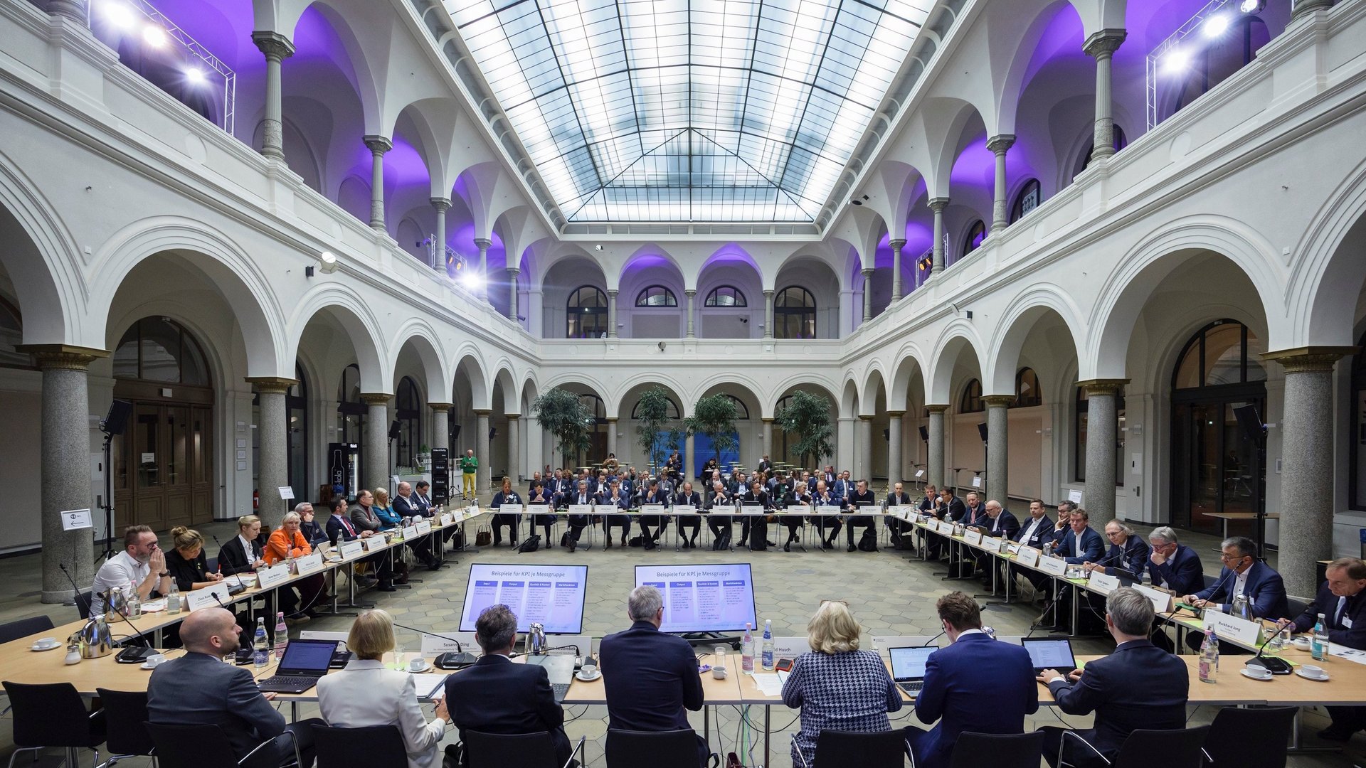 Blick in einen großen Saal mit Lichtauge und Arkaden, die den Raum umgeben. In einem Viereck sind Tische und Stühle aufgebaut, an denen die Anwesenden sitzen.