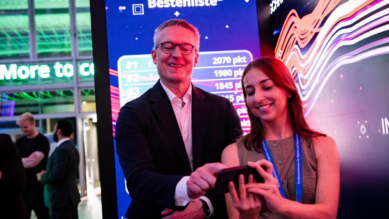 Minister Wildberger tippt auf ein Handy, das ihm eine Frau am Messestand reicht