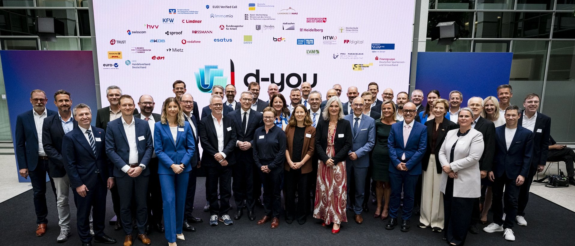 Eine große Gruppe von Personen steht für ein Gruppenfoto vor einer Präsentationsfläche mit dem d-you-Logo und zahlreichen Logos beteiligter Organisationen.