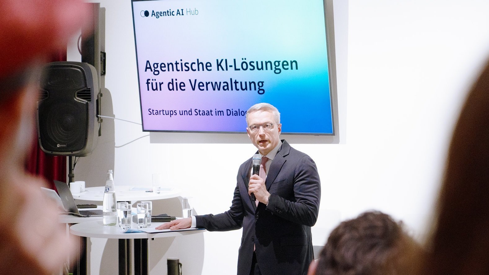 Ein Mann in einem dunklen Anzug steht auf einer Bühne und spricht in ein Mikrofon. Im Hintergrund ist ein großer Bildschirm zu sehen, auf dem der Text „Agentische KI-Lösungen für die Verwaltung – Startups und Staat im Dialog“ steht.