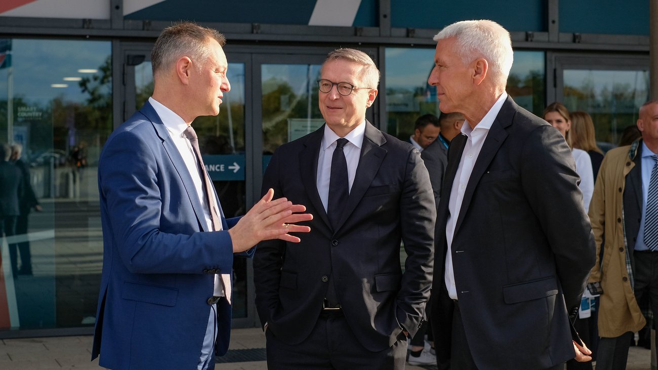 Drei Männer in Anzügen stehen vor einer Messehalle und Reden.