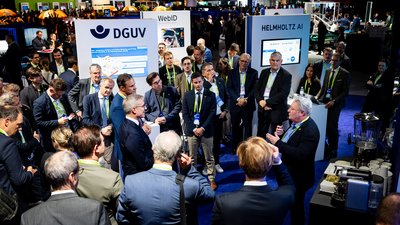 Präsentation an einem Messestand: viel Publikum versammelt sich um einen Redner. 