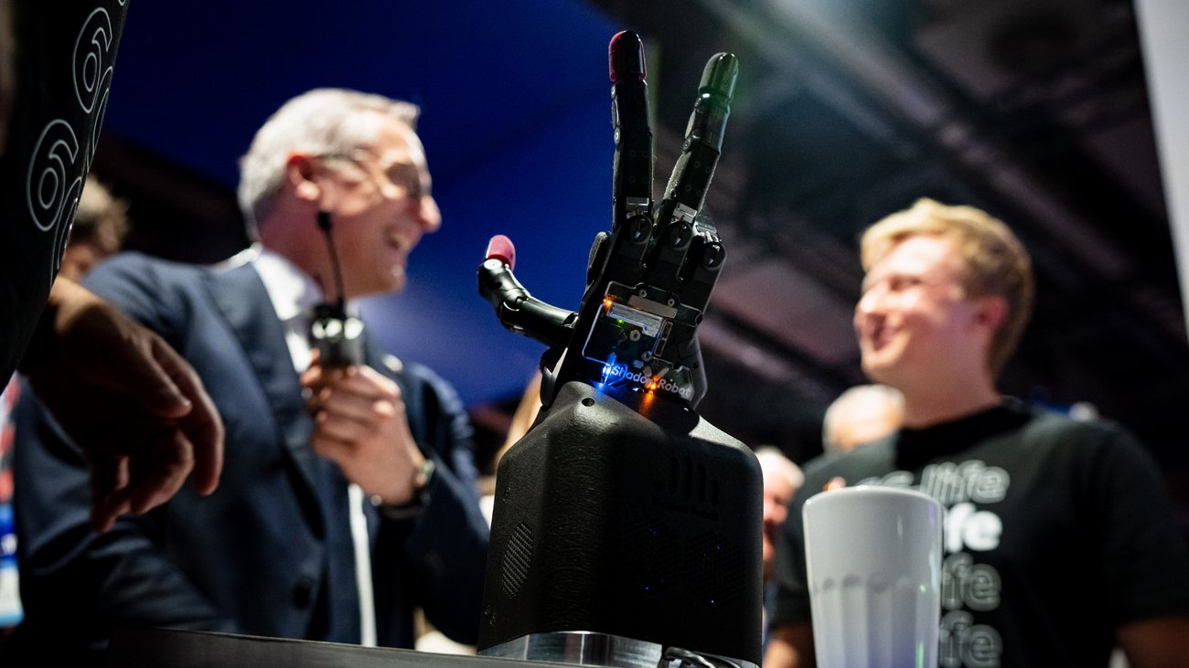 Eine Roboterhand scharf im Vordergrund,  im Hintergrund verschwommen zwei Personen. 
