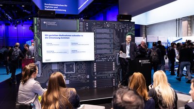 Bilder der Bühne am Messestand. Der Hintergrund ist in einem Design eines Mikrochips, darauf ein großer Monitor sowie Sitzwürfel im Vordergrund. 