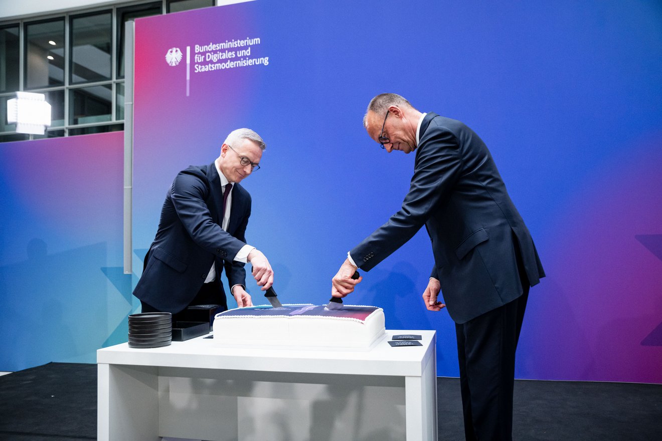 Karsten Wildberger und Friedrich Merz schneiden gemeinsam eine rechteckige Torte an. Im Hintergrund ist der Schriftzug "Bundesministerium für Digitales und Staatsmodernisierung" zu lesen.
