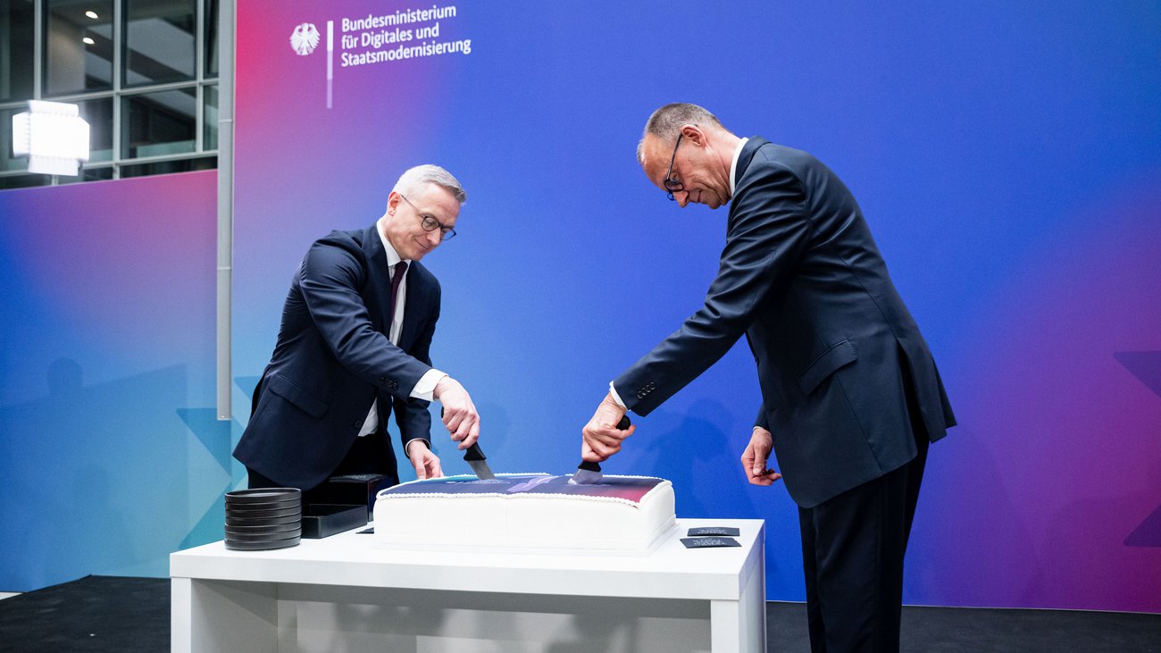 Karsten Wildberger und Friedrich Merz schneiden gemeinsam eine rechteckige Torte an. Im Hintergrund ist der Schriftzug "Bundesministerium für Digitales und Staatsmodernisierung" zu lesen.