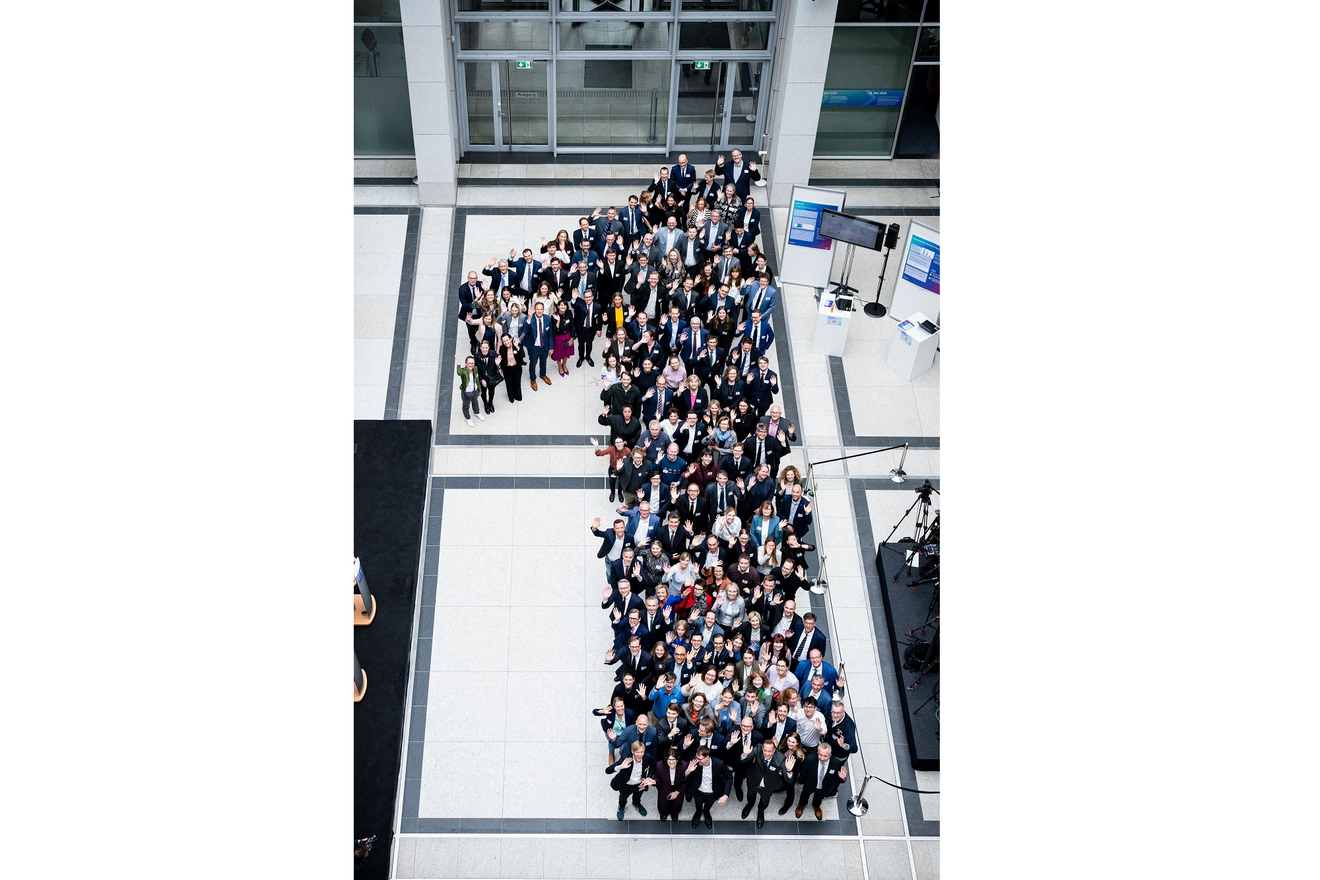 Ein großes Gruppenfoto in der Vogelperspektive, auf dem zahlreiche Menschen in einem modernen, hellen Foyer in der Form einer großen Ziffer „1“ aufgestellt sind. Die Personen tragen überwiegend formelle Business-Kleidung, blicken nach oben in die Kamera und winken teilweise.