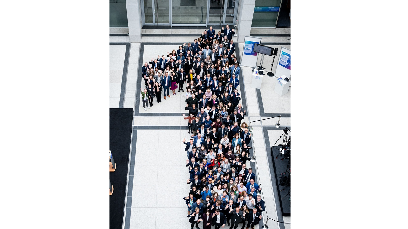 Ein großes Gruppenfoto in der Vogelperspektive, auf dem zahlreiche Menschen in einem modernen, hellen Foyer in der Form einer großen Ziffer „1“ aufgestellt sind. Die Personen tragen überwiegend formelle Business-Kleidung, blicken nach oben in die Kamera und winken teilweise.
