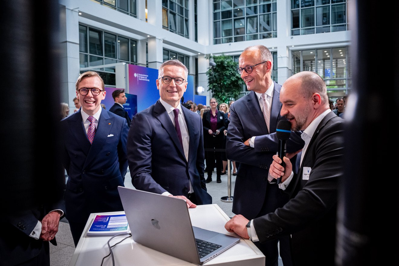 Ein Mann spricht in ein Mikrofon, auf einem Tisch vor ihm steht ein Laptop. Links neben ihm stehen Friedrich Merz, Karsten Wildberger und Philipp Amthor. Im Hintergrund sind weitere Personen zu sehen.