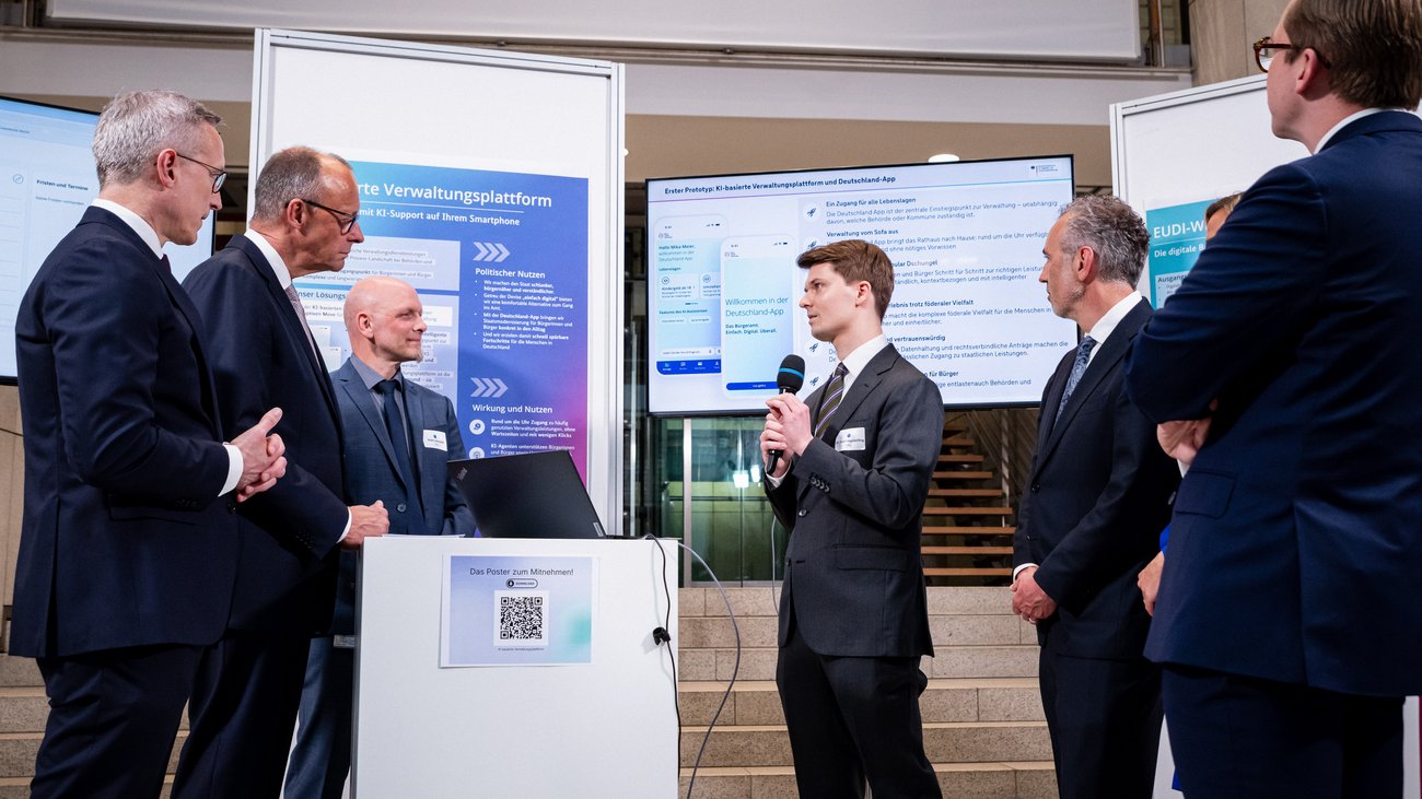 David Hagebölling spricht in ein Mikrofon und erläutert eine App. Um ihn herum stehen Friedrich Merz, Karsten Wildberger, Thomas Jarzombek und Philipp Amthor. Im Hintergrund sind Monitore und Whiteboards zu sehen.