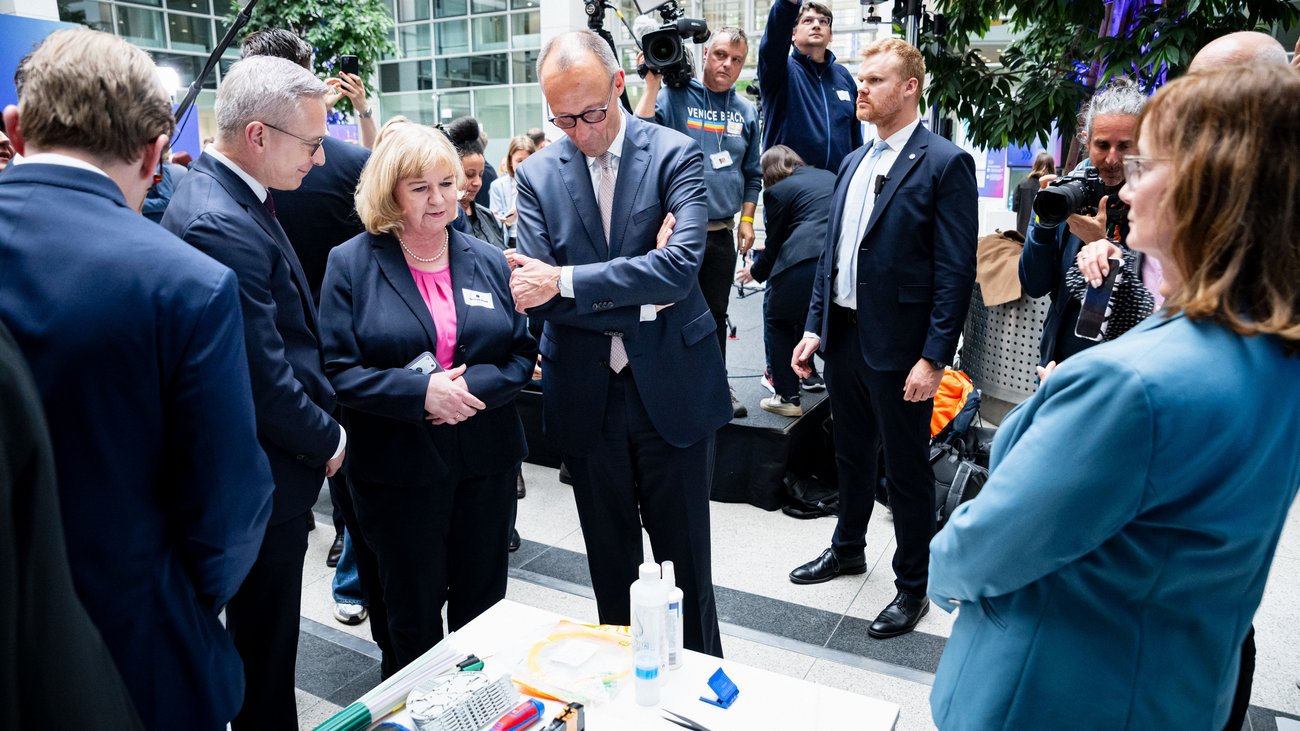 Getrud Husch, Friedrich Merz und Karsten Wildberger stehen vor einem Tisch und betrachten ein Exponat. Um sie herum stehen weitere Personen.