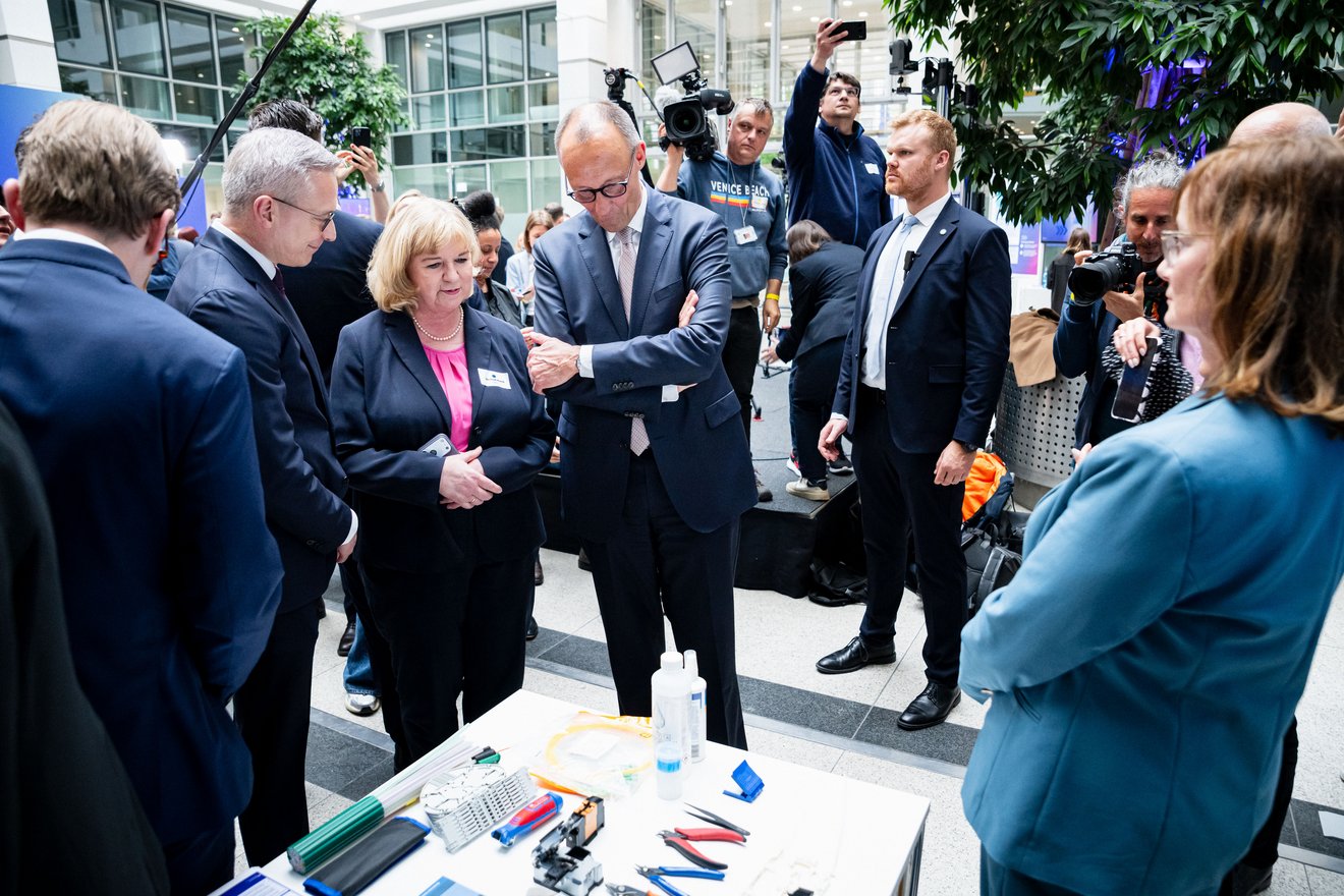 Getrud Husch, Friedrich Merz und Karsten Wildberger stehen vor einem Tisch und betrachten ein Exponat. Um sie herum stehen weitere Personen.