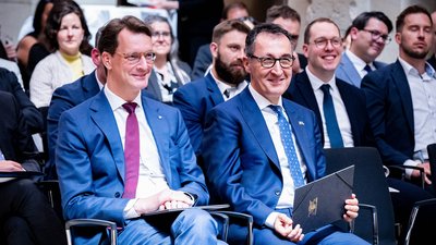 Hendrik Wüst und Cem Özdemir sitzen lächelnd in einem Publikum