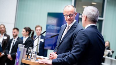 Friedrich Merz steht an einem Rednerpult und blickt zu Karsten Wildberger, der am Rednerpult neben ihm steht. Im Hintergrund sind weitere Personen und ein Bildschirm zu sehen.