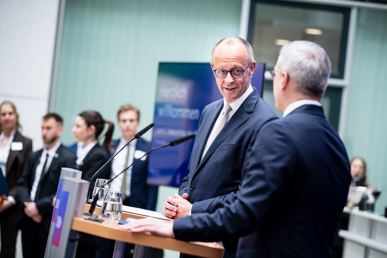 Friedrich Merz steht an einem Rednerpult und blickt zu Karsten Wildberger, der am Rednerpult neben ihm steht. Im Hintergrund sind weitere Personen und ein Bildschirm zu sehen.