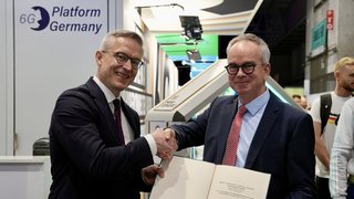 Zwei Männer geben sich die Hand. Eine der Personen hält ein geöffnetes Buch in der Hand. Im Hintergrund ist ein Messestand mit der Aufschrift "6G Platform Germany" zu sehen.