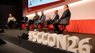 Bühnenszene schräg von der Seite: 5 Personen sitzen auf einer Bühne, im Vordergrund die Buchstaben #SCCON26.