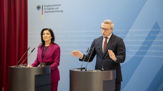 Zwei Personen stehen an Rednerpulten vor einer hellblauen Wand mit dem Logo und Schriftzug des „Bundesministeriums für Digitales und Staatsmodernisierung“. Links steht eine Frau in einem fuchsiafarbenen Hosenanzug, rechts ein Mann im dunklen Anzug mit Brille, der während des Sprechens gestikuliert. Im Hintergrund ist ein schwerer roter Vorhang zu sehen.